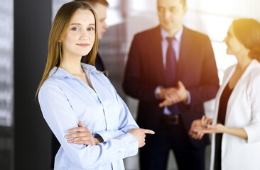 Young blonde businesswoman dressed in blouse is standing with arms crossed in a sunny modern office. Successful young business people