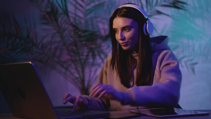 Female working with headphones, a tablet and a laptop on a colorful neon light background. Close-up.