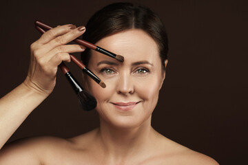 Poster - Beautiful middle aged woman with a makeup brushes on brown background