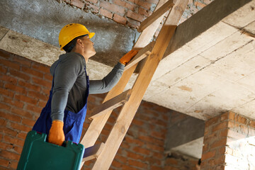 Wall Mural - From the ground up. Young male builder in blue overalls and hard hat climbing up the ladder, holding toolbox while working at construction site