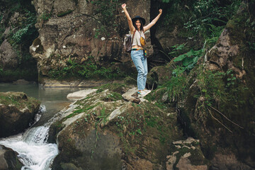 Wall Mural - Happy woman traveler with backpack standing on rocks of river in mountains,  holding hands up