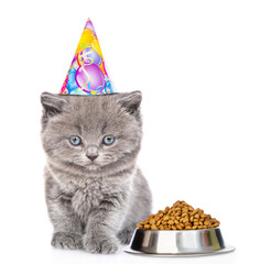 Poster - Cute kitten wearing party cap sits with bowl of dry food for pet and looks at camera. isolated on white background