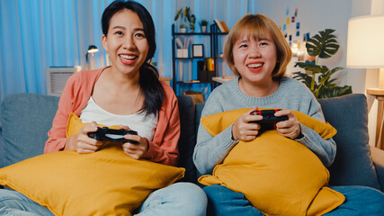 Sticker - Lesbian LGBTQ women couple play video game at home. Young Asia lady using wireless controller having funny happy moment on sofa in living room at night. They have great and fun time celebrate holiday.