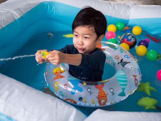 Asian cute child boy playing water splash in blue bowl with funny face and wet hair in rubber band. Kid having happy moment in summer. Concept of family activity, songkran festival at home.