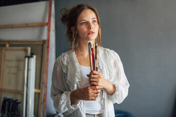 Wall Mural - Candid portrait of an artist female posing in her art studio with paintbrushes in hands. A professional young woman painter searching for an imagination in her workshop.