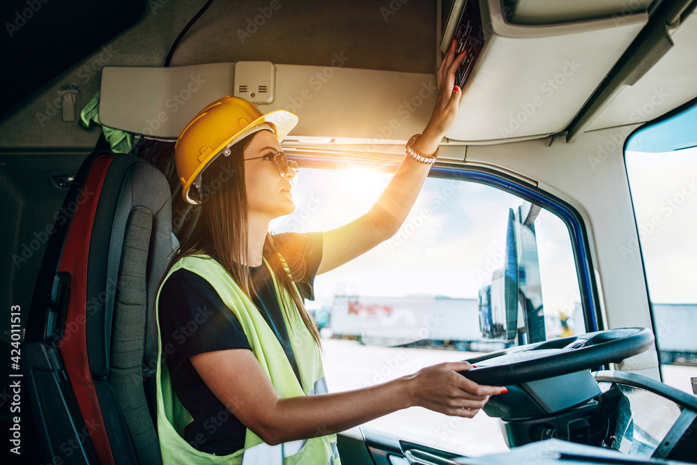 Portrait of beautiful young woman professional truck driver with protective yellow helmet sitting and driving a big truck. Inside of vehicle. People and industrial transportation concept. - obrazy, fototapety, plakaty 