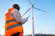 A woman engineer, holding a tablet and working at the wind turbine farm in the field in the orange vesta