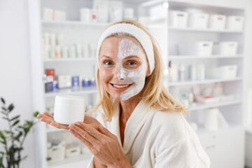 Senior female with cosmetic mask on half face at beauty salon. Woman smiles and holds bootle of facial mask.