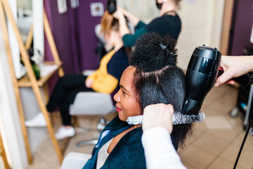 A hairdresser dries a Black woman's hair in a modern hair salon. Beauty/fashion concept. Caucasian client in a background.