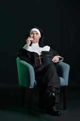 Wall Mural - Senior nun with cigar and Bible sitting in armchair on dark background