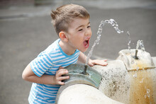 A Little Joyful Boy Drinks From A Fountain And Catches A Stream Of Water With His Mouth On A Sunny Day In The City.