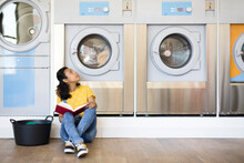 Hispanic Woman Sitting On The Floor While Waiting For Clothes Washing In The Self-service Laundry. Space For Text.