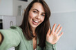 © Drobot Dean - Young happy woman waving hand while taking selfie photo at home kitchen