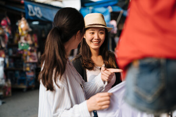 Wall Mural - two girl friends together spend time travel together in summer bangkok thailand. Two beautiful woman make purchases in outdoor clothing store vendor in market. female point showing sister shopping