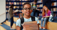 Middle Eastern Boy Holding Stack Of Books Against Multi Colored Bookshelf In Library.