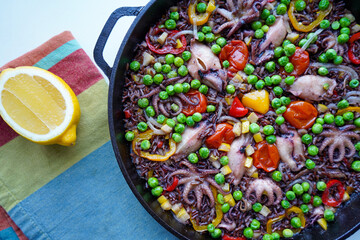 Wall Mural - Close Up of Colorful Spanish Seafood Paella Dish with Octopus and Squid Served in Shallow Pan with Black Handles with Fresh Lemon in Background