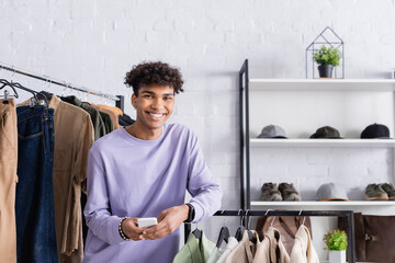 Cheerful african american showroom owner holding smartphone near clothes