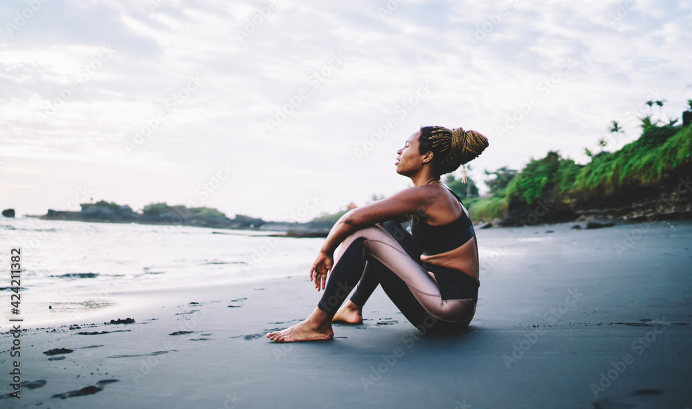 African American female runner resting at black sandy beach enjoying morning time for training body muscles, casual woman in tracksuit keeping healthy lifestyle getting energy on workout near sea - obrazy, fototapety, plakaty 
