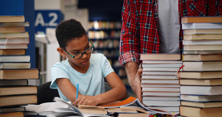 Wall Mural - Librarian bringing stack of books to teen african schoolboy in library