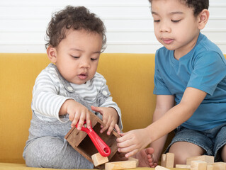 A mixed-race Asian-African boy about 11 months old sits on a sofa and playing a wooden toy with his brother together in the house. Funny educational for children concept..