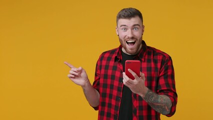 Surprised shocked bearded tattooed young man 20s in red checkered shirt isolated on yellow background in studio. People lifestyle concept. Using mobile cell phone pointing index finger aside say wow