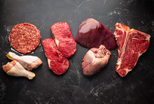 Selection Of Assorted Raw Meat Food For Zero Carb Carnivore Diet: Uncooked Beef Steak, Ground Meat Patty, Heart, Liver And Chicken Legs On Black Stone Background From Above 