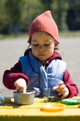 Little girl play with set of children's toy colorful dishes, cones and chestnutson on the bench. Kid toy utensil for girls game. Early spring