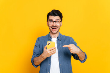 happy joyful caucasian stylish guy with glasses, holding smartphone in hand and pointing at it finge