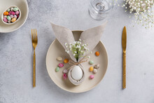 Easter Table Setting With Egg In Napkin In The Form Of An Easter Bunny With Ears, With Small White Flowers On Ceramic Plate And Cutlery. Easter Table Decorations. Top View