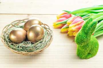 Wall Mural - Easter eggs basket. Golden egg in basket with spring tulips, white feathers on wooden background in Happy Easter decoration. Traditional decoration in sun light.