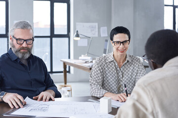 Wall Mural - Mature business couple listening to young architect while sitting at the table with blueprints during business interview at office