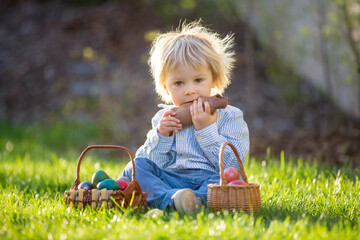 Little toddler boy, eating chocolate bunny in garden on sunset, easter eggs around him