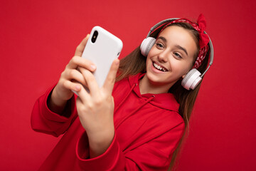 Closeup photo of smiling Positive attractive brunette girl wearing red hoodie isolated on red background holding and using smartphone communicating online wearing white wireless headphones listening