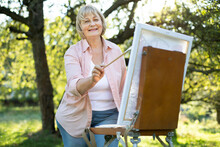 Portrait Of Positive Talented Middle Aged Woman Artist Standing In Front Of Easel Outdoors In The Green Park, Holding Brush, Working On A Picture. Creativity, Inspiration, Art And Painting Concept