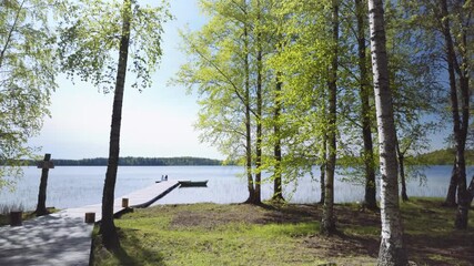 Sticker - Walk by the lake among the trees on a sunny spring day. Boats with oars moored to the shore. Large stones covered with moss lie on the green grass. Latvia