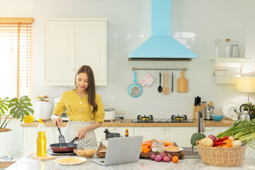 A beautiful Asian girl makes breakfast by looking at how to make it with her laptop computer with a happy face. At her own house