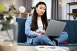© sofiko14 - Close up portrait of pretty confident purposeful Asian Korean woman, working remotely from home, sitting ar cozy living room on sofa in lotus position and typing on laptop pc