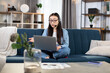© sofiko14 - Confident young woman in headset sitting on couch and having video conference on laptop. Female freelancer leading working meeting online while staying at home.
