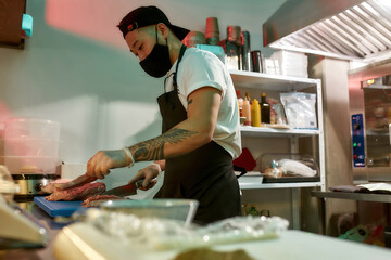 Wall Mural - Young male cook wearing protective mask and gloves cutting raw pork meat on a plastic board while working in commercial kitchen