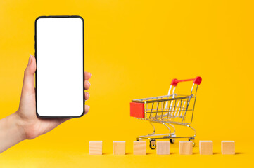 An empty cart and wooden blocks. On the left, a woman's hand holds a smartphone. Mock up. Yellow background. Copy space. The concept of economy, crisis and shopping