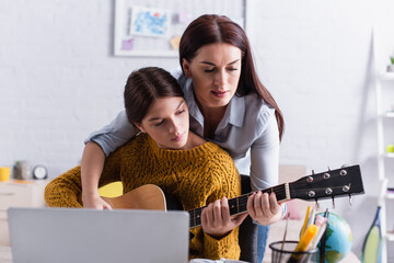 Wall Mural - mother assisting teenage girl playing acoustic guitar near laptop