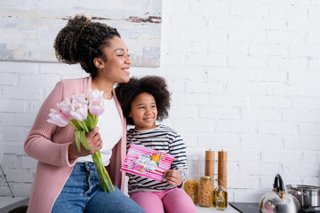 Happy African American woman and girl with happy mothers day card smiling and looking away in kitchen