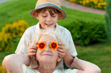 Wall Mural - Boy puts grandmother on the eyes of chamomile. Family holiday .Laughing grandson with his beloved grandmother spend leisure time outdoors. An elderly woman and a grandchild. Summer spring time