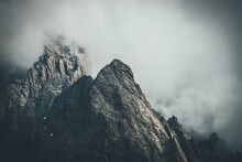 Dark Atmospheric Surreal Landscape With Dark Rocky Mountain Top In Low Clouds In Gray Cloudy Sky. Gray Low Cloud On High Pinnacle. High Black Rock With Snow In Low Clouds. Surrealist Gloomy Mountains.