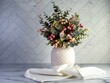 White ceramic vase with raised dots on it filled with a green and pink leafy plant with little peach and pink round balls sitting on a marble counter with a white towel and a herringbone tile.