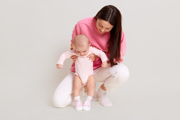 Wall Mural - Young dark haired beautiful mother wearing casual sweater and pants with infant baby, mommy teaching daughter to walk and looking at kid, posing isolated on white background.