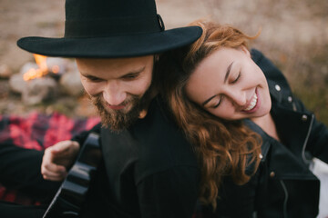 Wall Mural - Happy couple in love relaxing sitting by bonfire near lake, playing ukulele and hugging.