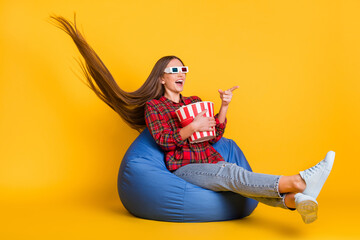 Wall Mural - Full size photo of optimistic brunette long hairdo lady hold pop corn wear spectacles shirt jeans sit on bag isolated on yellow background