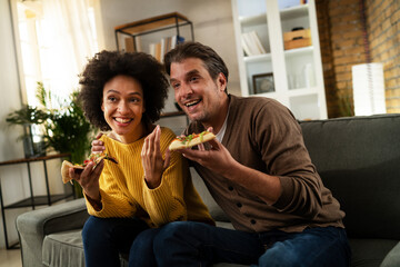 Wall Mural - Cheerful young couple sitting on sofa at home. Happy woman and man eating pizza while watching a movie.