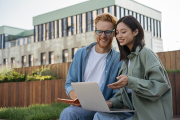 Two smiling students studying, learning, exam preparation sitting in university campus. Education concept. Colleagues using laptop computer, communication, talking, working together outdoors